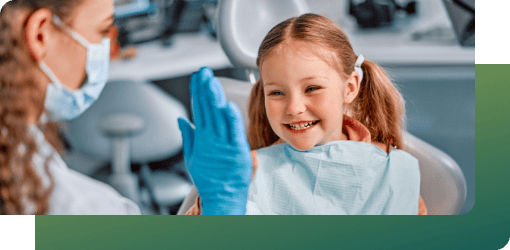 A dental professional checking a smiling young girl’s teeth and gums at a children’s dental care clinic for her first visit.